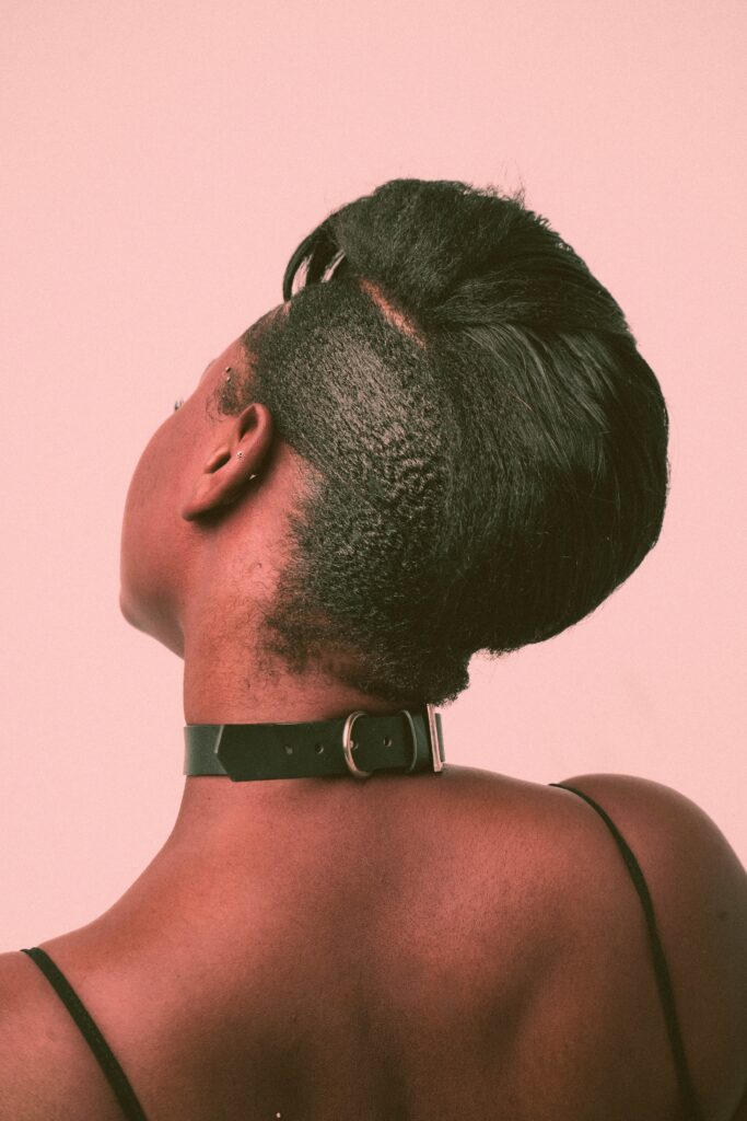 The back of a black woman with a short edgy haircut highlights the leather collar she wears around her neck